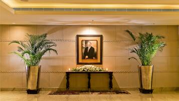 A lobby with a marble floor, a wooden table with a framed portrait, two potted plants, and a lit candle.