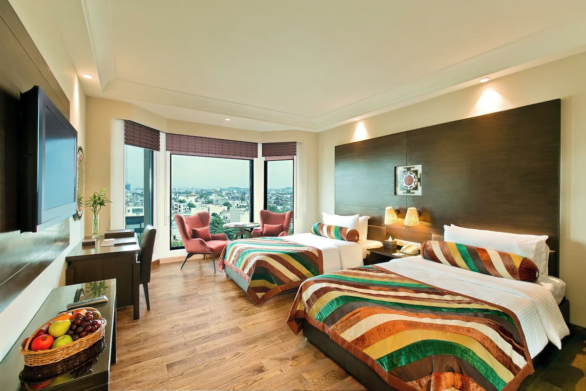 Ein Hotelzimmer mit zwei Betten, einem Fernseher, einem Schreibtisch mit Obstkorb und Blick auf die Stadt.