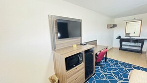 Une chambre d’hôtel dotée d’un téléviseur à écran plat fixé au mur, d’un bureau en bois avec un four à micro-ondes, d’une chaise rouge et d’une salle de bain avec un miroir et un lavabo.
