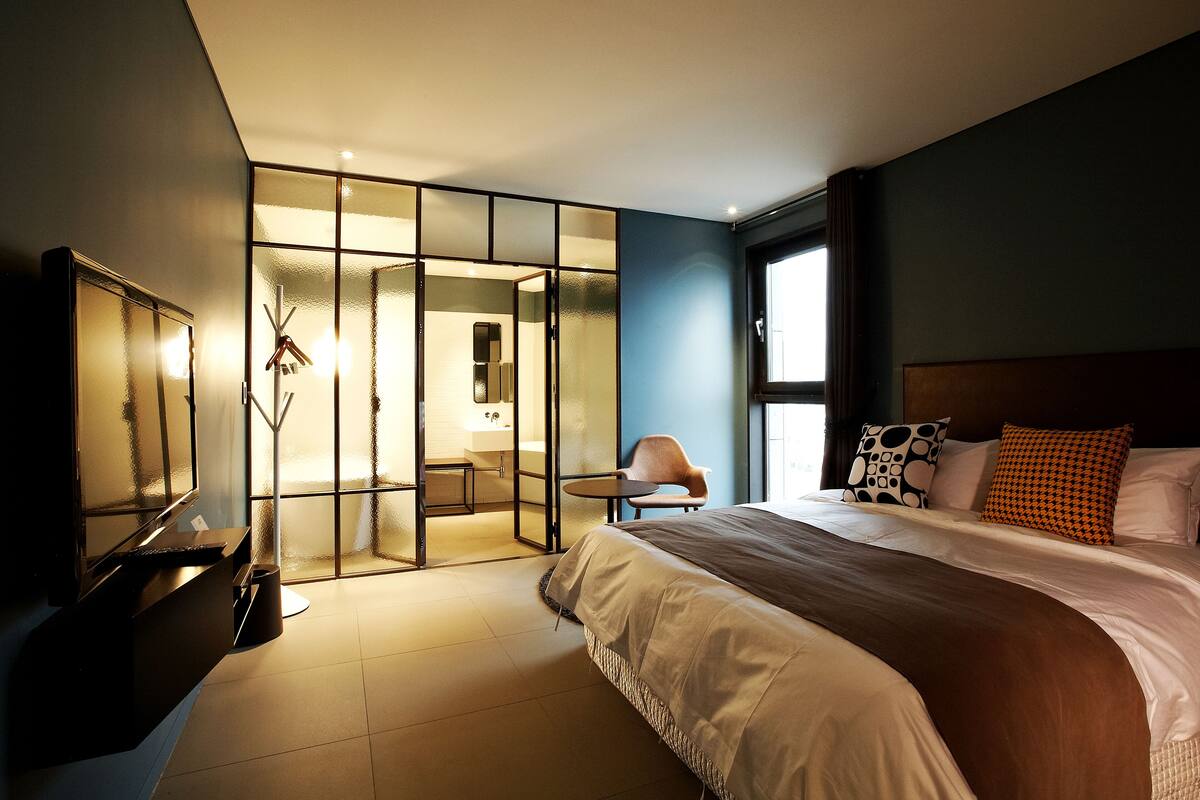 A modern hotel room with a large bed, a desk, and a glass partition separating the room from a bathroom.