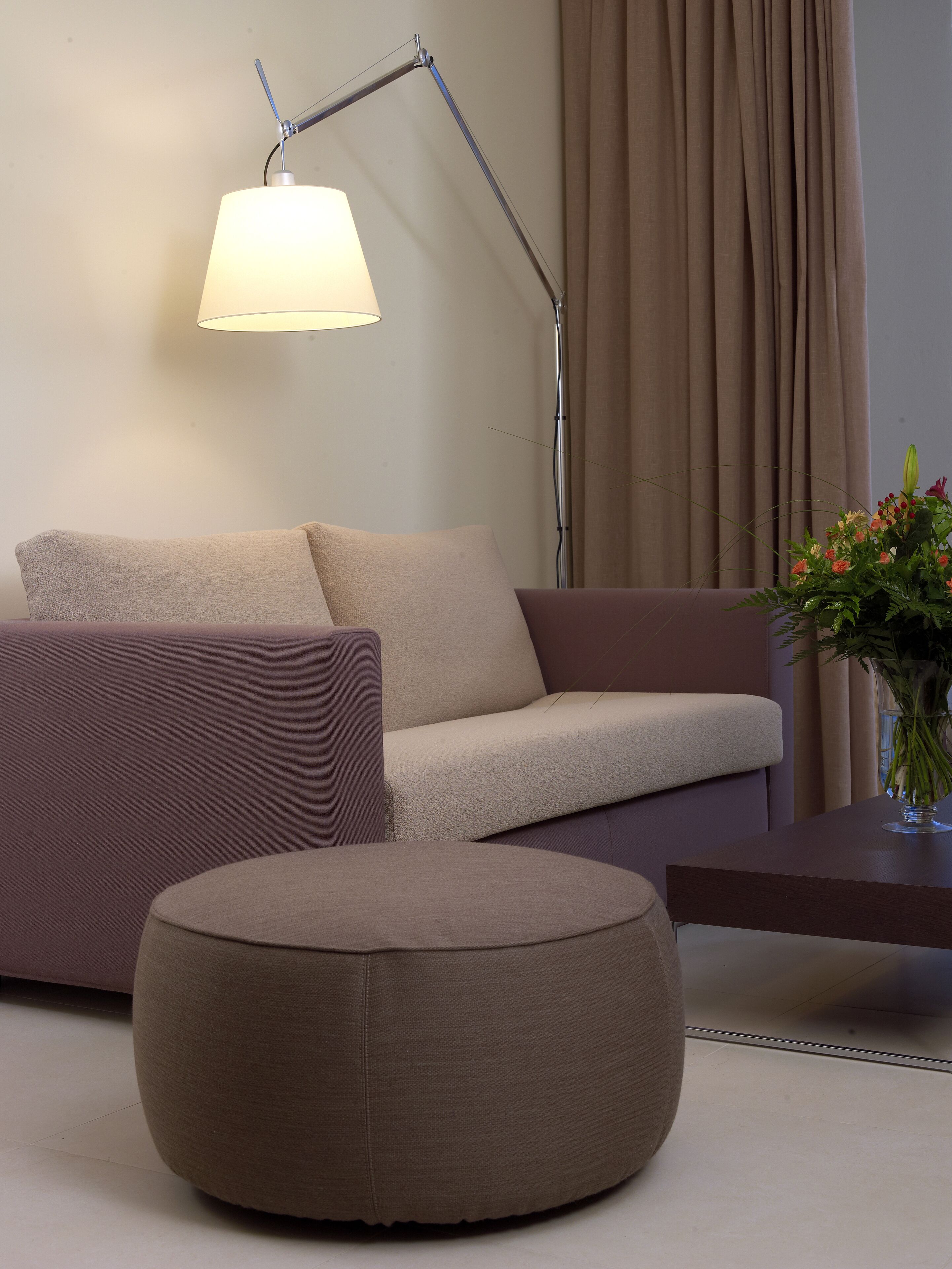Una sala de estar moderna con un sofá, una otomana, una lámpara de pie y un florero con flores sobre una mesa.