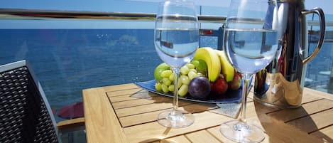 A table with a fruit platter, two wine glasses, and a silver pitcher on a boat deck with a view of the sea.