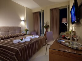 Habitación de hotel con cama, escritorio con un florero, silla, televisor y mesa de vidrio con copas de vino y fruta.