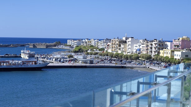 A waterfront promenade with a glass railing overlooking the sea, a row of outdoor seating, and a view of the cityscape in the background. - El Greco Hotel (Ierapetra)