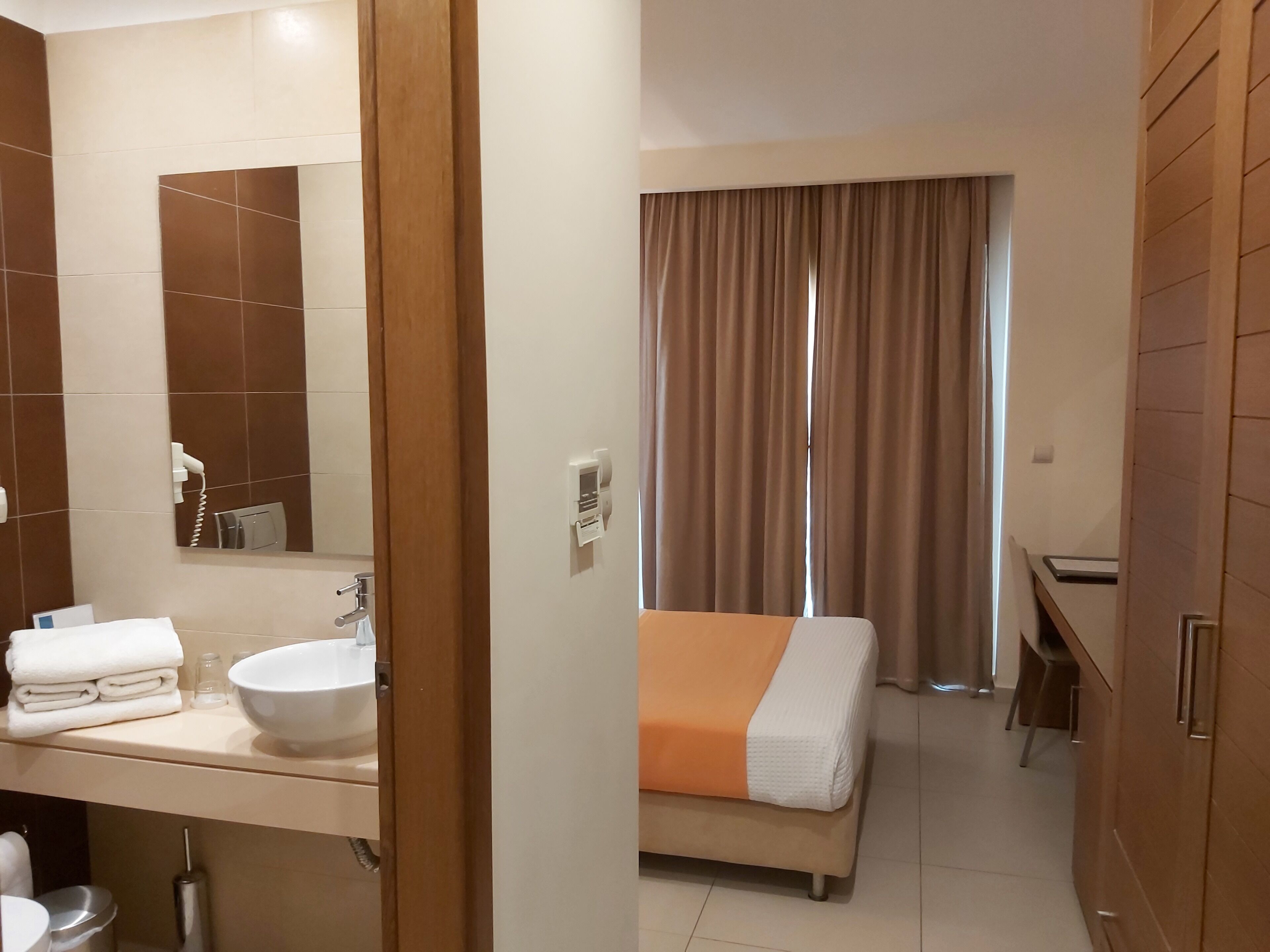 Hotelkamer met badkamer, een bed met oranje dekbed, spiegel, wastafel en raam met gordijnen.