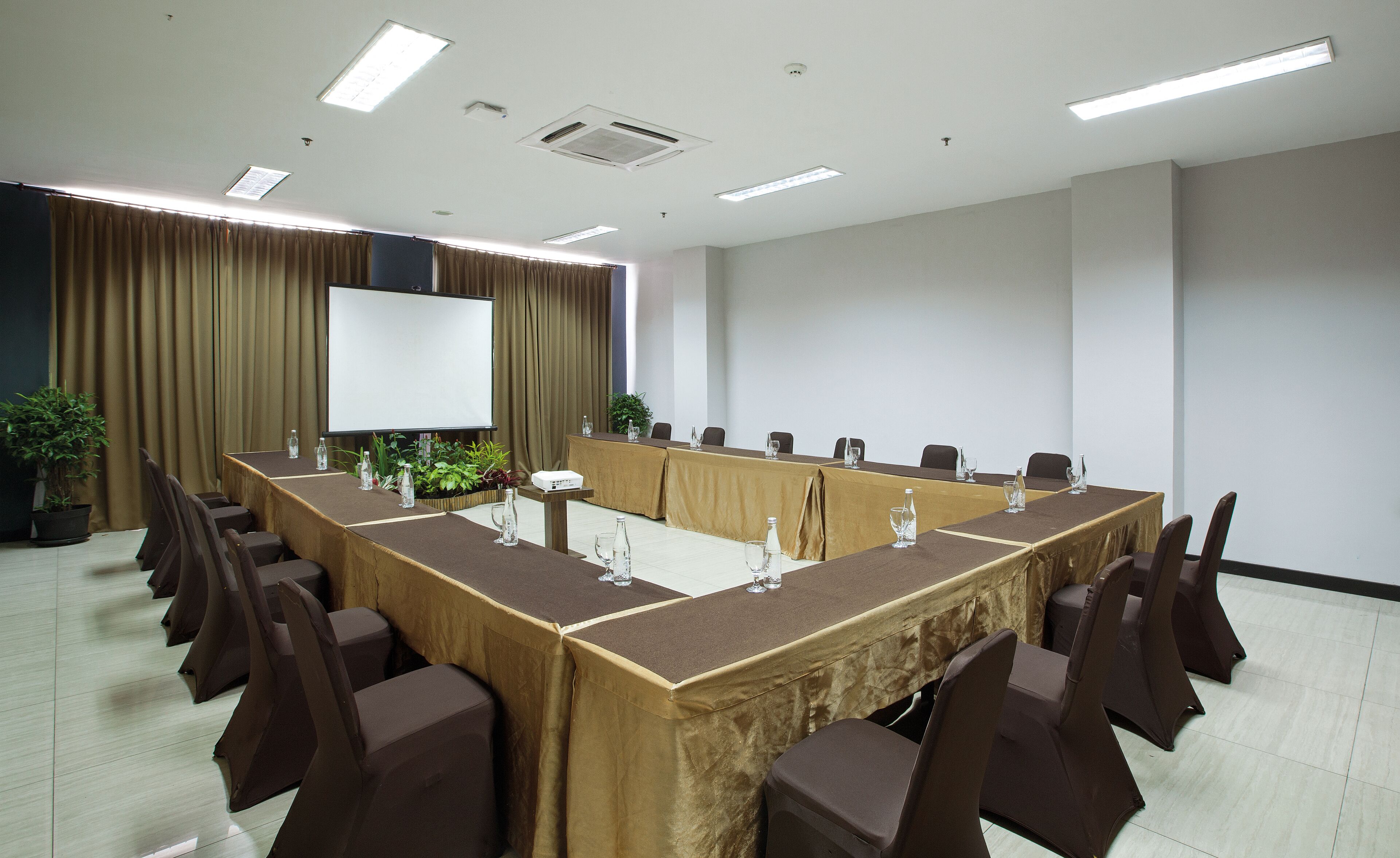 A conference room with a long table, chairs, a projector screen, and a potted plant.
