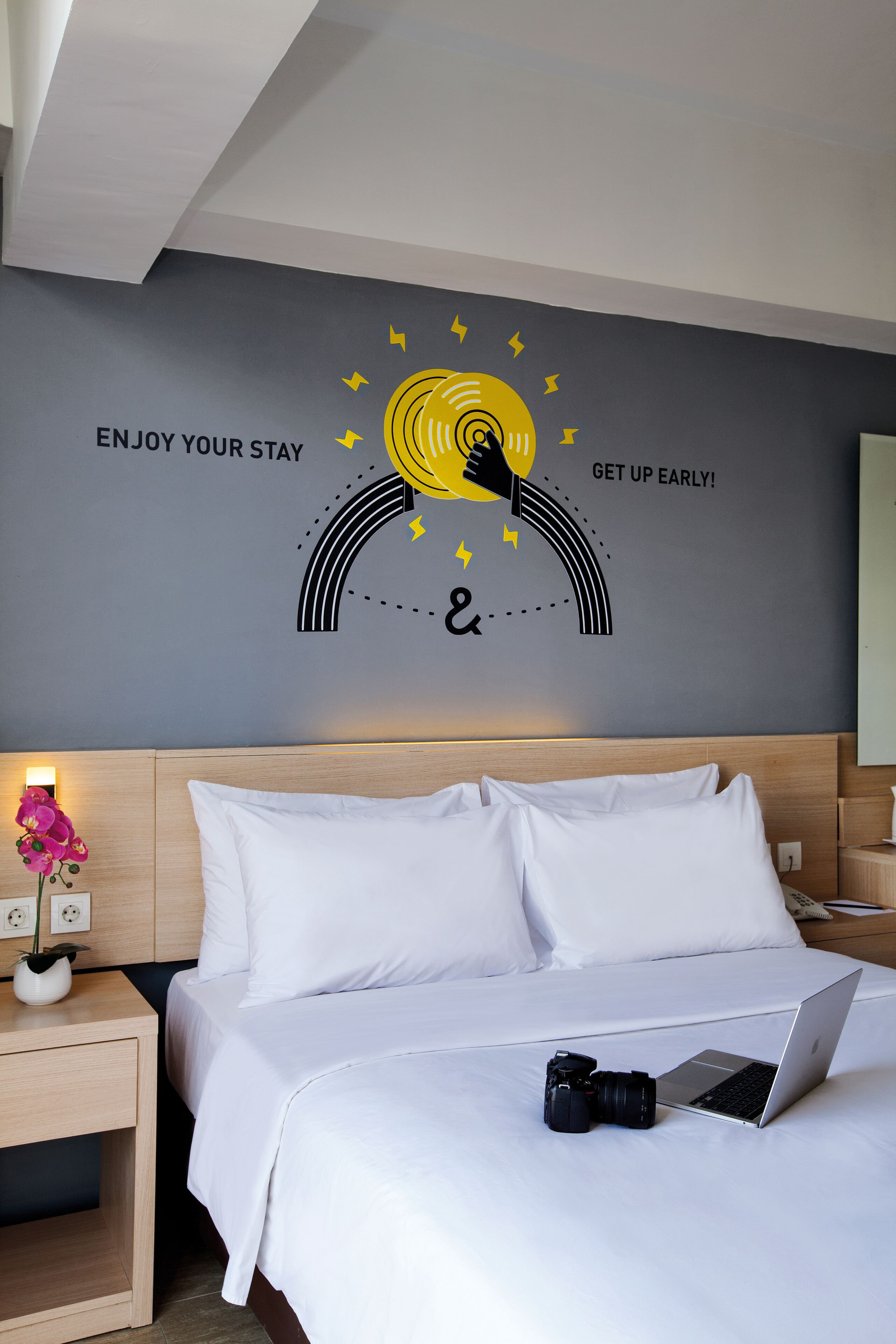 A hotel room with a bed, a nightstand with a lamp, and a wall decal with motivational text.