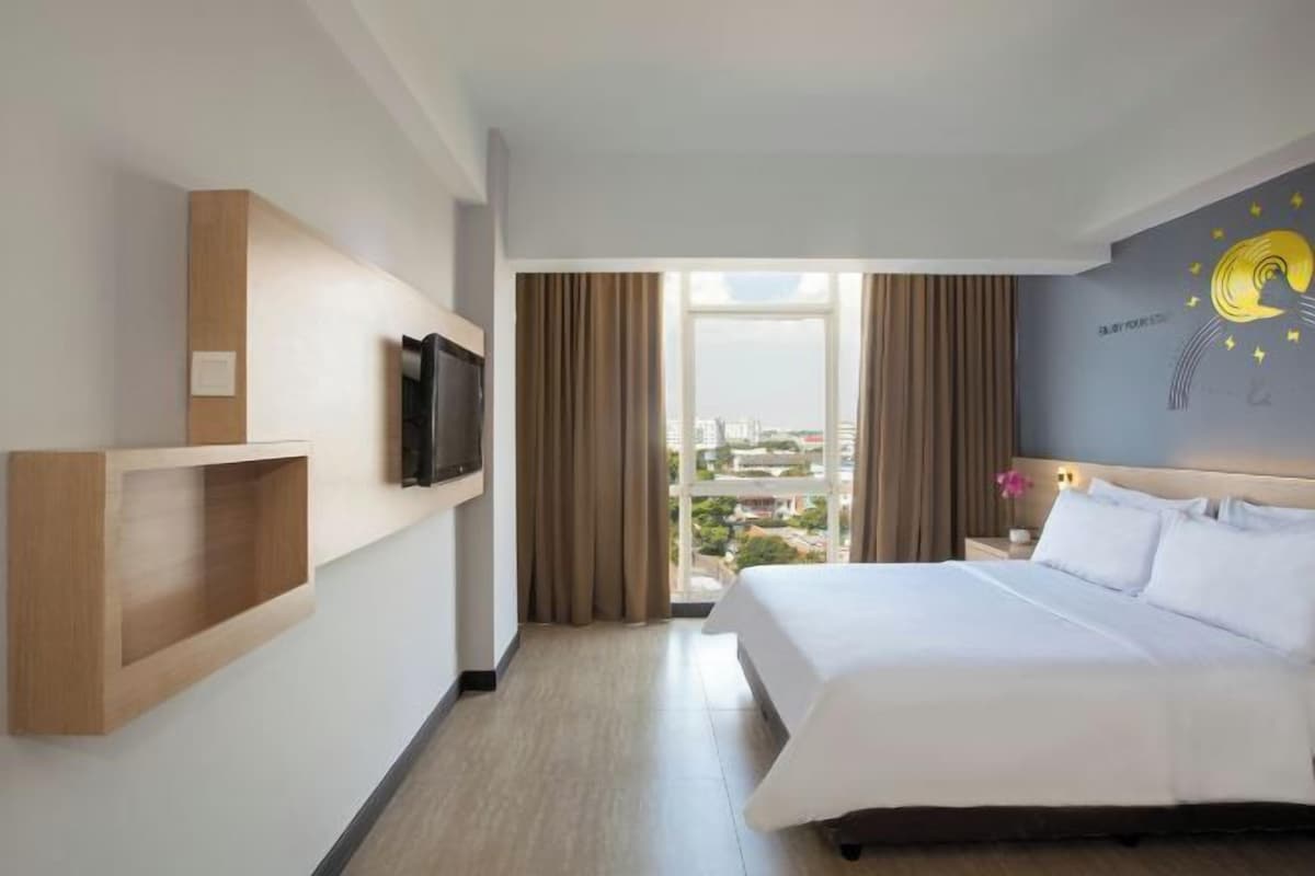 Une chambre d’hôtel avec un grand lit, un téléviseur à écran plat fixé au mur, une étagère en bois et une vue sur la ville par la fenêtre.