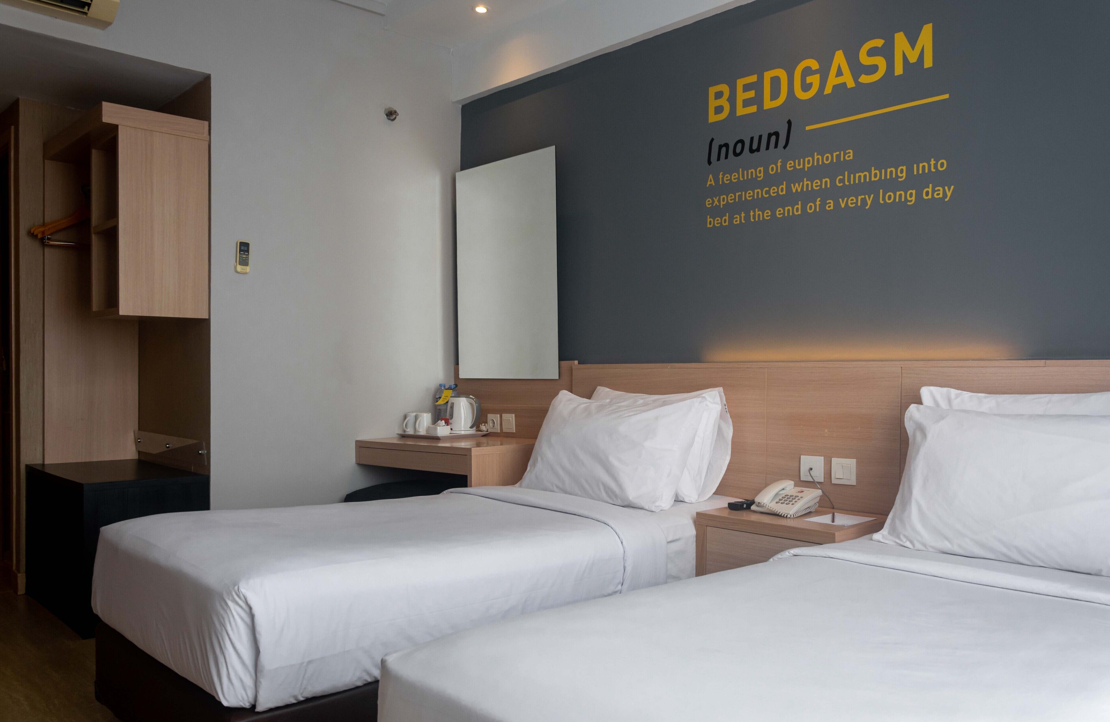 A hotel room with two beds, a bedside table with a lamp, a mirror, and a wall with a definition of 'bedgasm'.