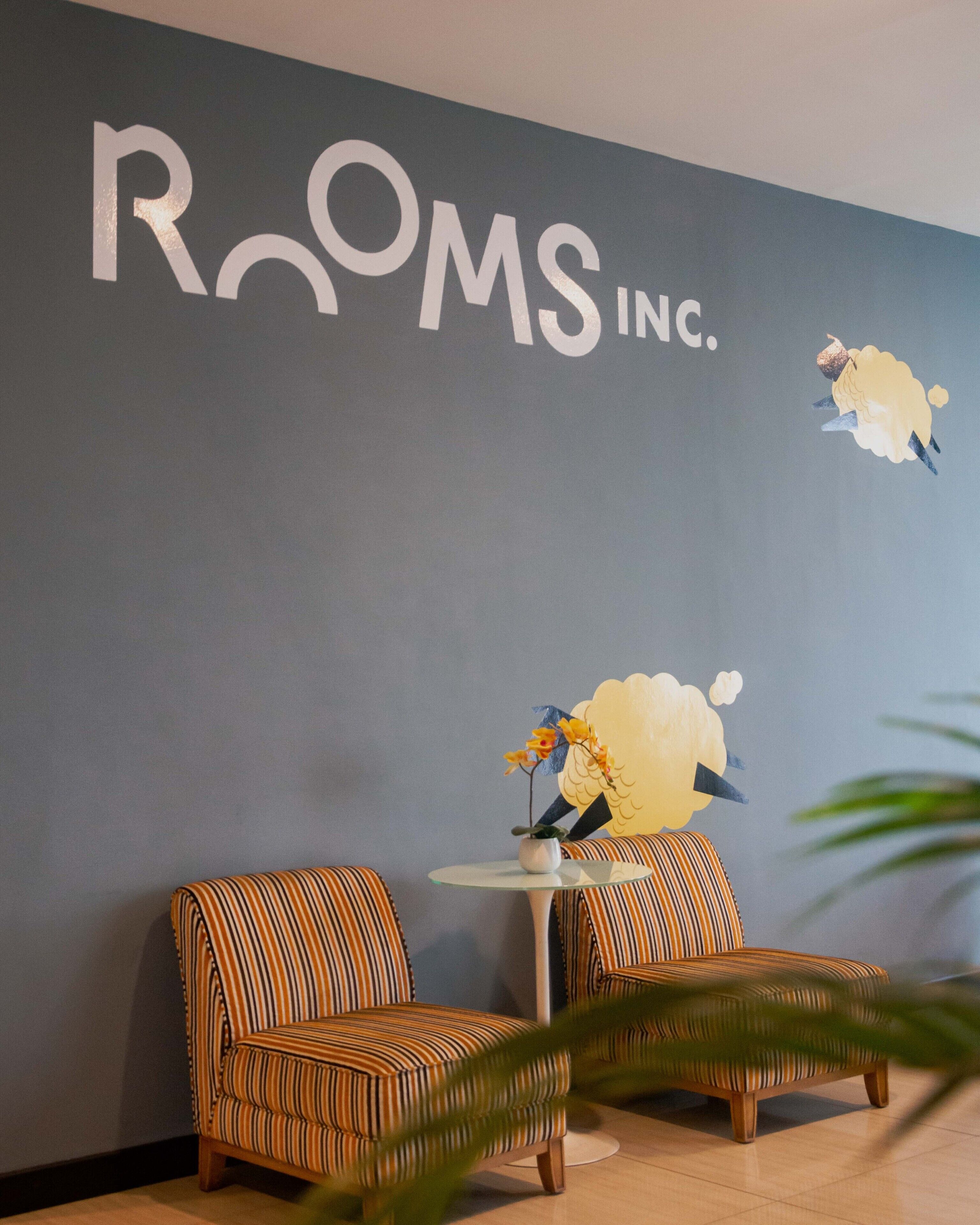 A room with striped chairs, a small table with a vase of flowers, and a wall with the text "ROOMS INC."
