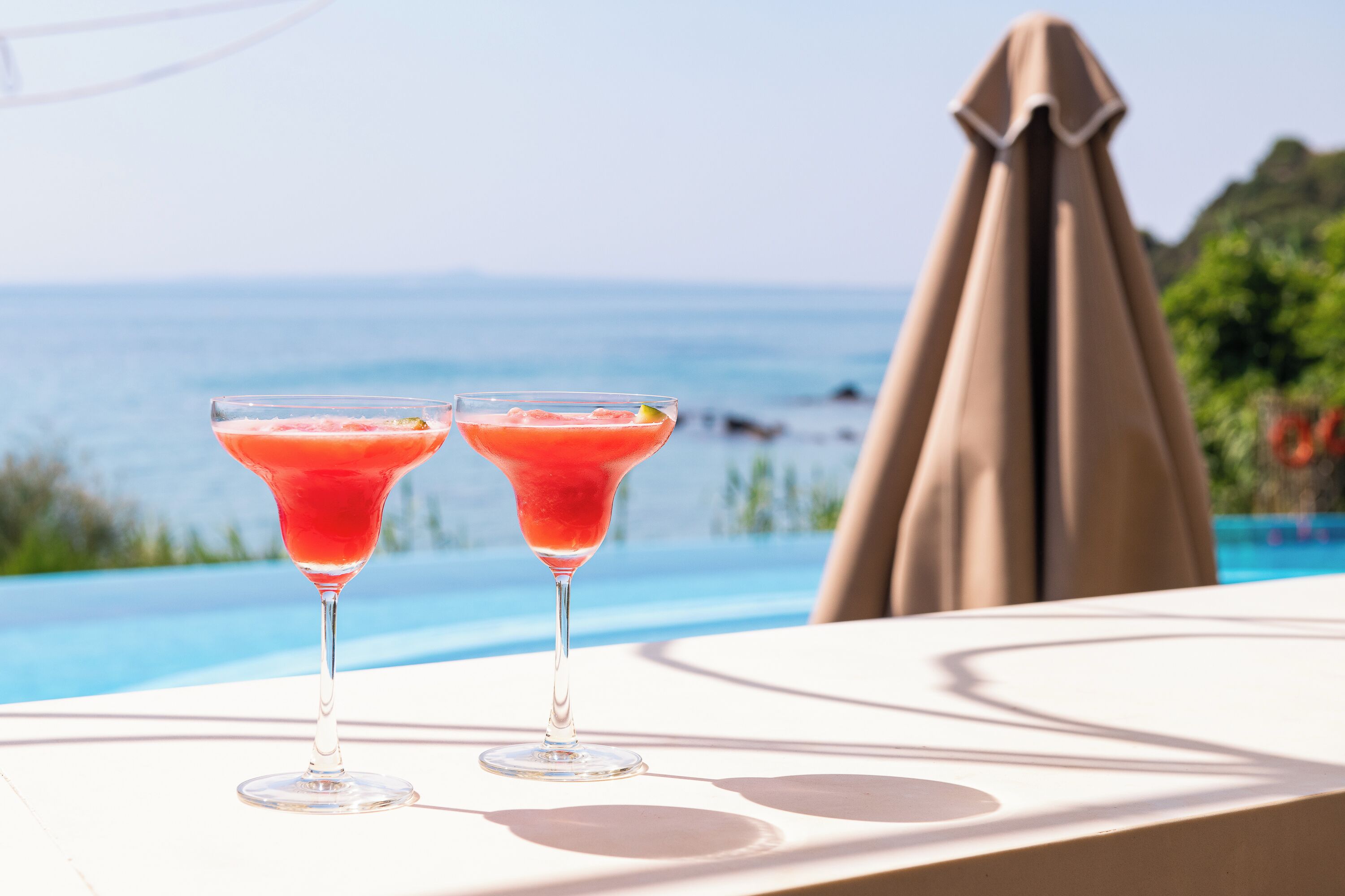 Two cocktails on a table by a pool with a view of the ocean.