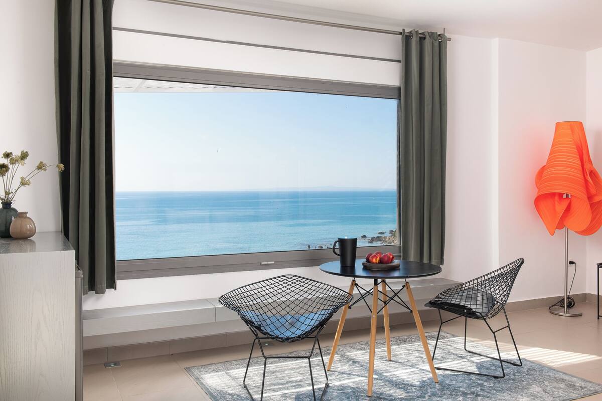 A room with a large window overlooking the sea, a round table with two chairs, a vase with dried flowers, and a bright orange lamp.