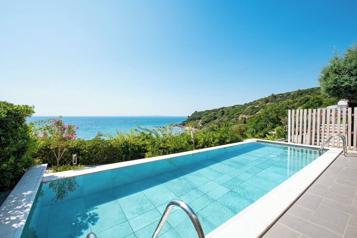 A swimming pool with a clear view of the sea, surrounded by greenery and a white fence.
