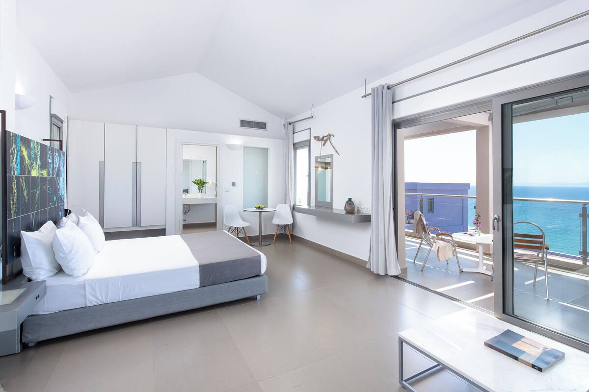 A modern hotel room with a bed, a desk, and a balcony with a view of the sea.