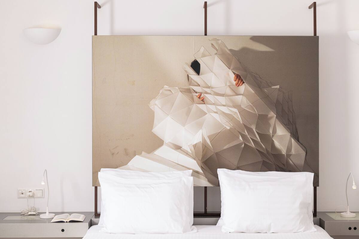 A modern bedroom with a geometric abstract painting on the wall, a bedside table with a lamp, and a neatly made bed with pillows.