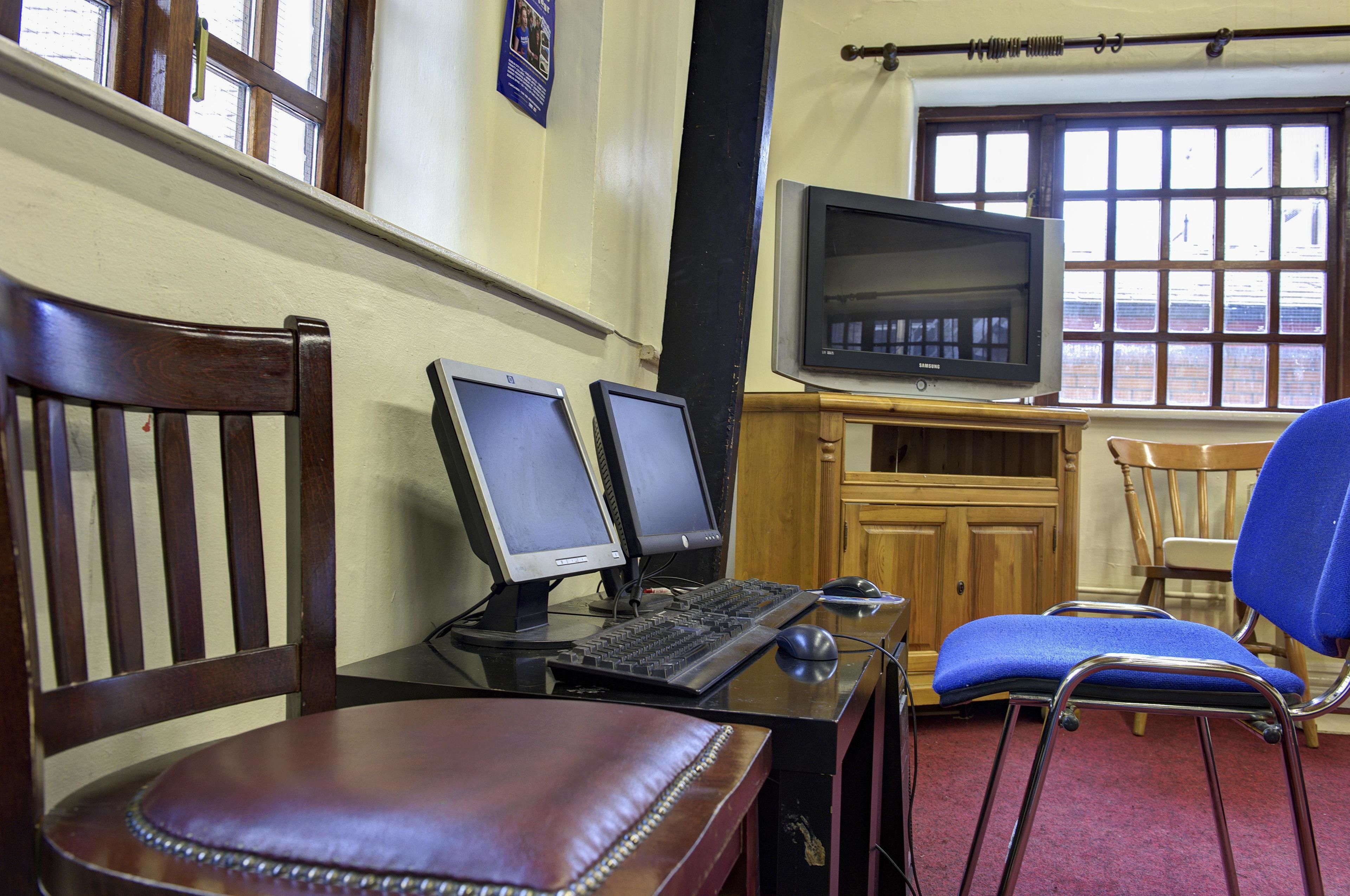 A room with two computer monitors, a keyboard, a mouse, and a television on a wooden desk. There are two chairs, one with a blue cushion and another with a brown leather seat. The room has wooden walls and a window with blinds.