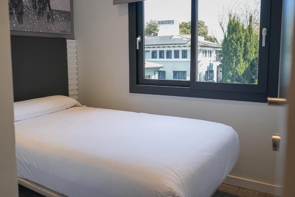 Yhden hengen sänky hotellihuoneessa, jonka ikkunasta avautuu näkymä rakennuksiin ja puihin.