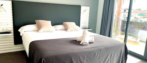 A hotel room with a large bed, a dark headboard, and a view of a balcony with palm trees.