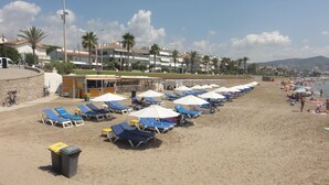 A beach with rows of sun loungers and umbrellas, a trash bin, and a building with a sloped roof in the background. - Hotel Sitges (Sitges)