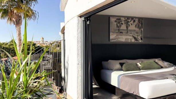 A modern bedroom with a bed, pillows, and a view of the cityscape. - Hotel Sitges (Sitges)