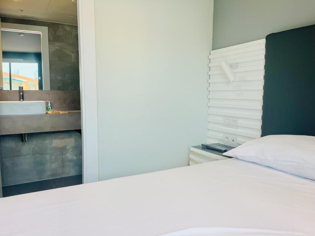 A modern hotel room with a bed, a white bedside table with a lamp, a large mirror, and a bathroom with a sink and towel rack.