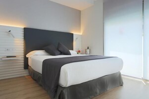 A modern bedroom with a large bed, a dark headboard, bedside tables, and a window with natural light.