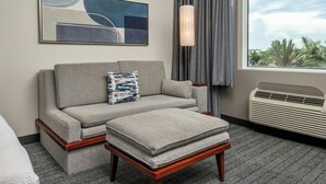 A hotel room with a sofa, an ottoman, a wall-mounted artwork, a floor lamp, a window with curtains, and a wall-mounted heater.