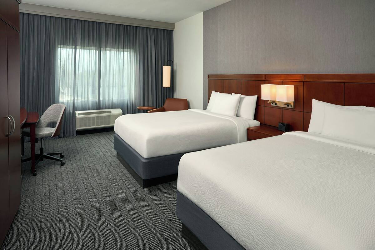 A hotel room with two beds, a desk, a chair, a window with curtains, and a wall-mounted light fixture.