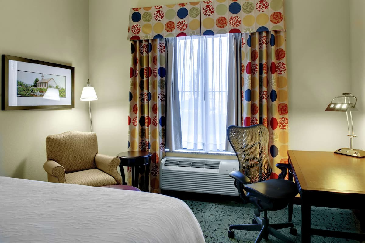 Une chambre d’hôtel avec un lit, un bureau, une chaise, une lampe et une fenêtre avec des rideaux.