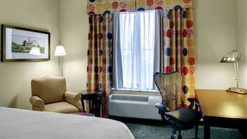 Quarto de hotel com cama, escrivaninha, cadeira, luminária e janela com cortinas.