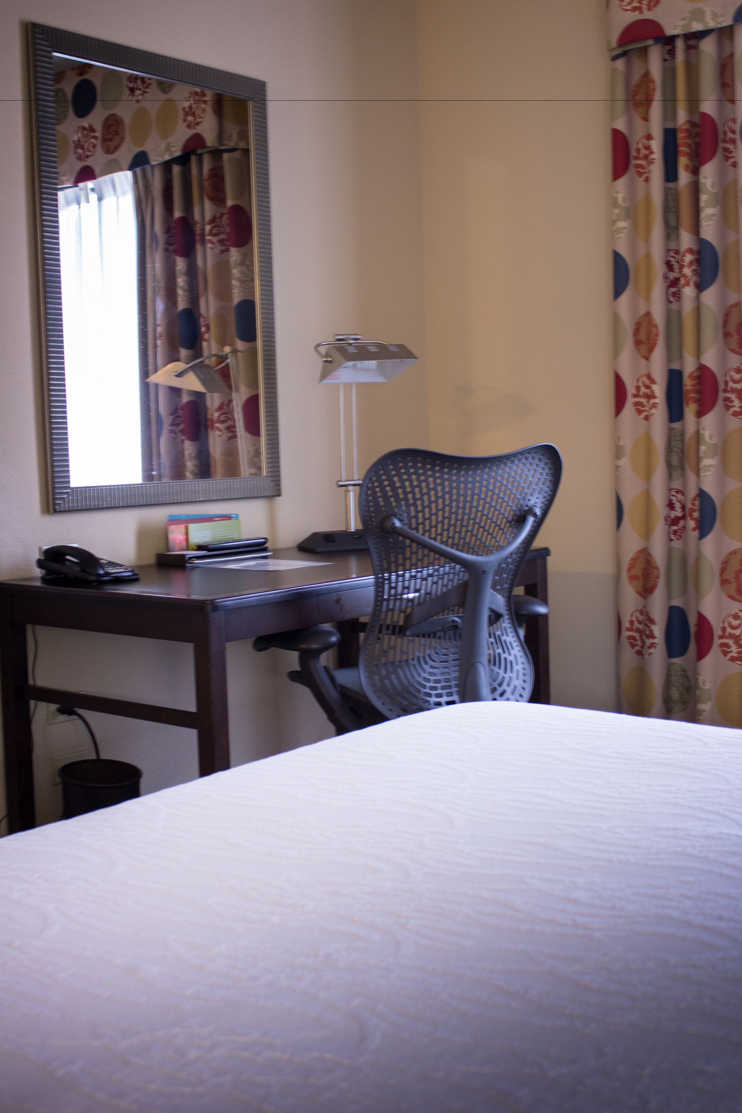 Habitación de hotel con una cama, un escritorio con teléfono, una silla, una lámpara y un espejo.