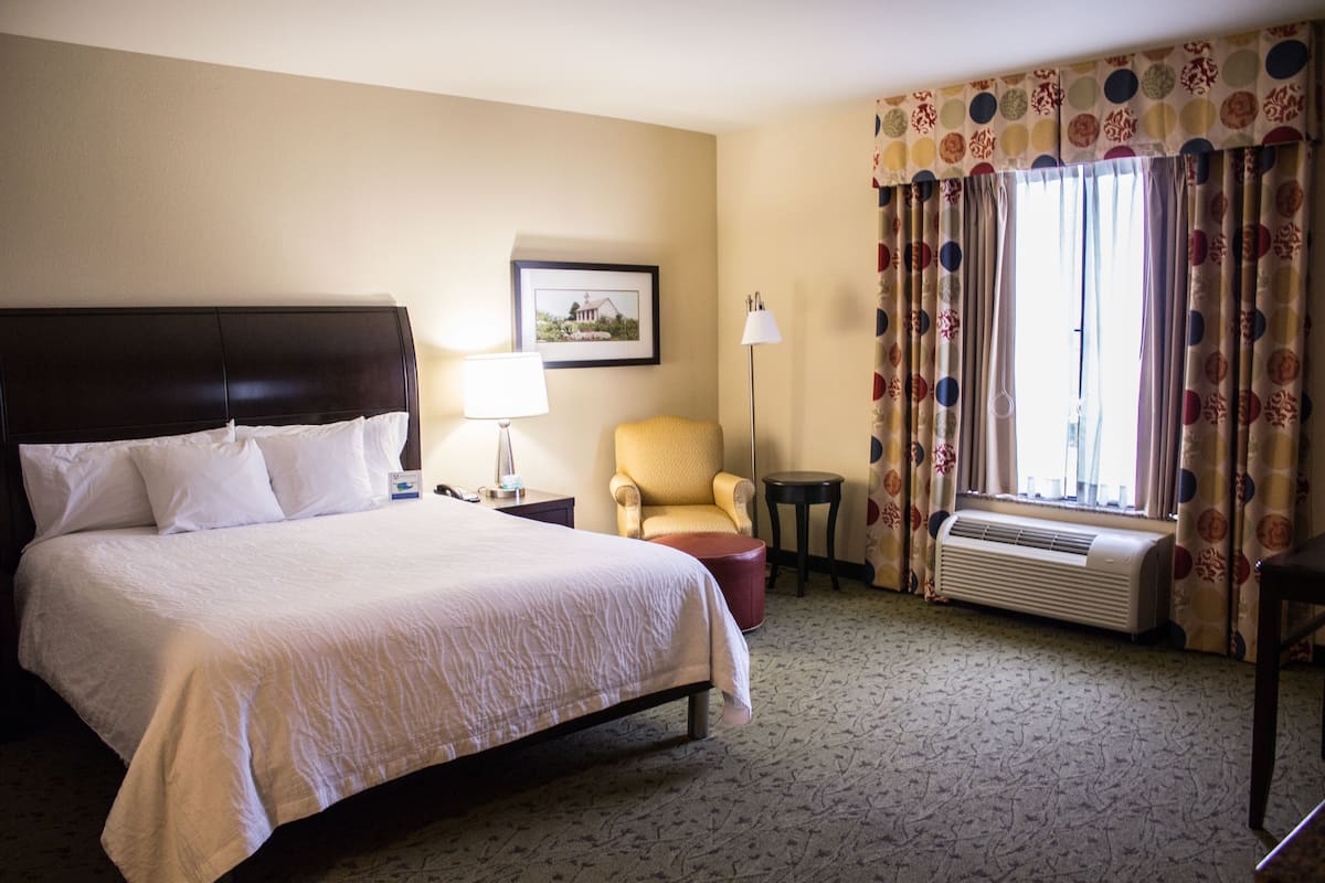 Une chambre d’hôtel avec un grand lit, une chaise, une petite table, une lampe, un tableau au mur et une fenêtre avec des rideaux.