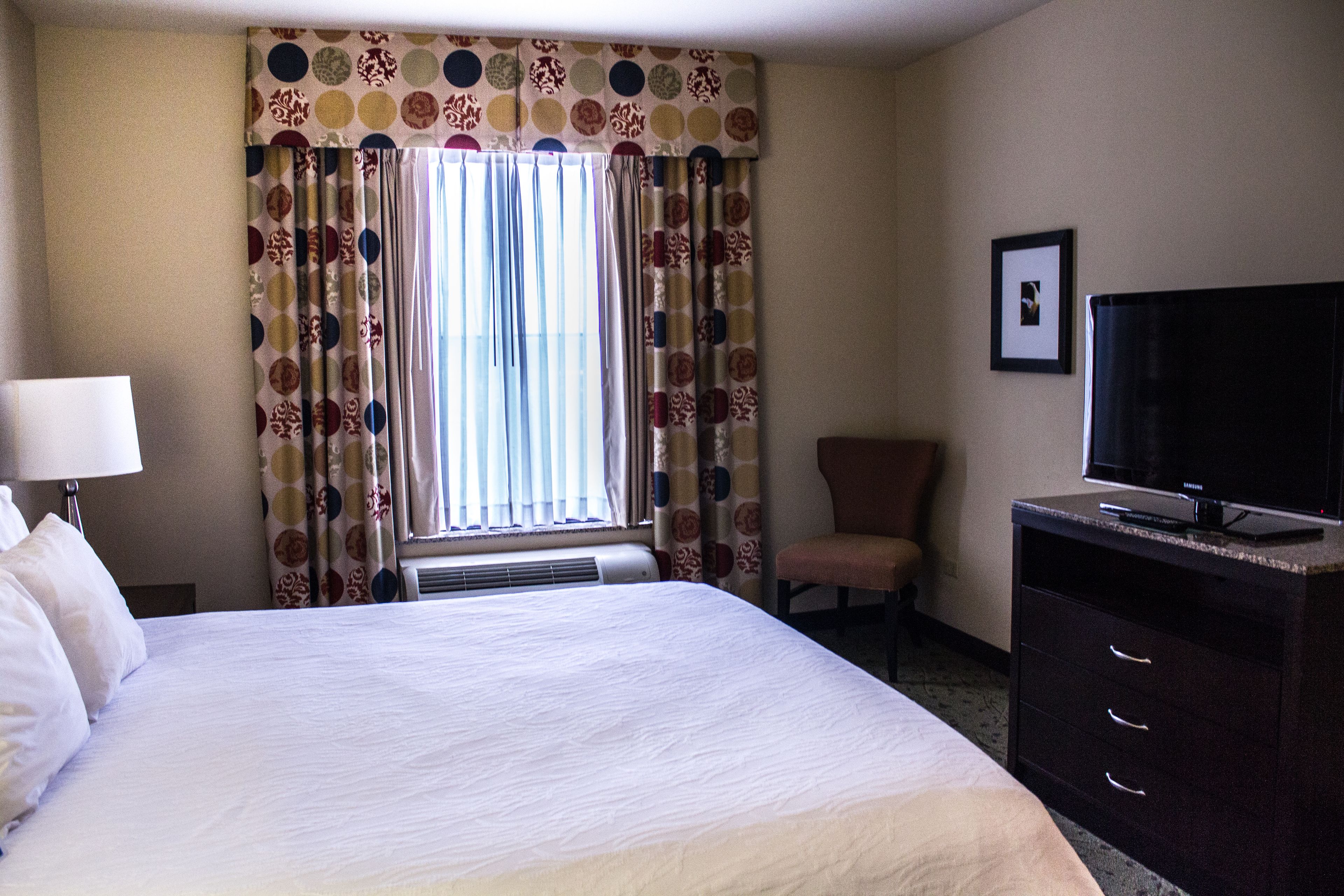 A hotel room with a large bed, a television, a chair, and a desk with drawers.