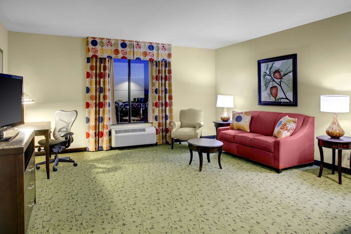 Une chambre d’hôtel équipée d’un canapé, d’un fauteuil, d’un bureau et d’une grande fenêtre avec des rideaux.