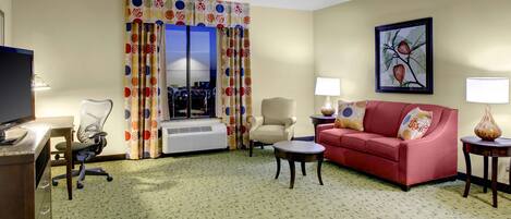 Quarto de hotel com sofá, poltrona, escrivaninha e uma janela grande com cortinas.