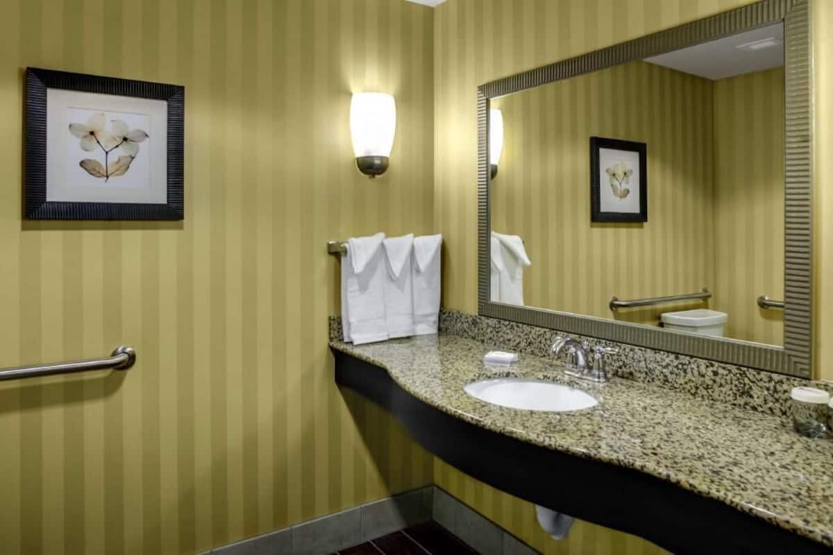 Une salle de bain avec un papier peint à rayures, un lavabo encastré dans un plan de travail en granit, un grand miroir et des œuvres d’art encadrées accrochées au mur.