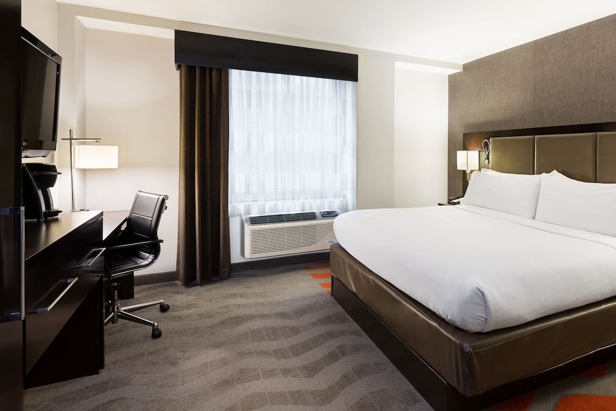 A hotel room with a large bed, a desk with a chair, a desk lamp, and a window with curtains.