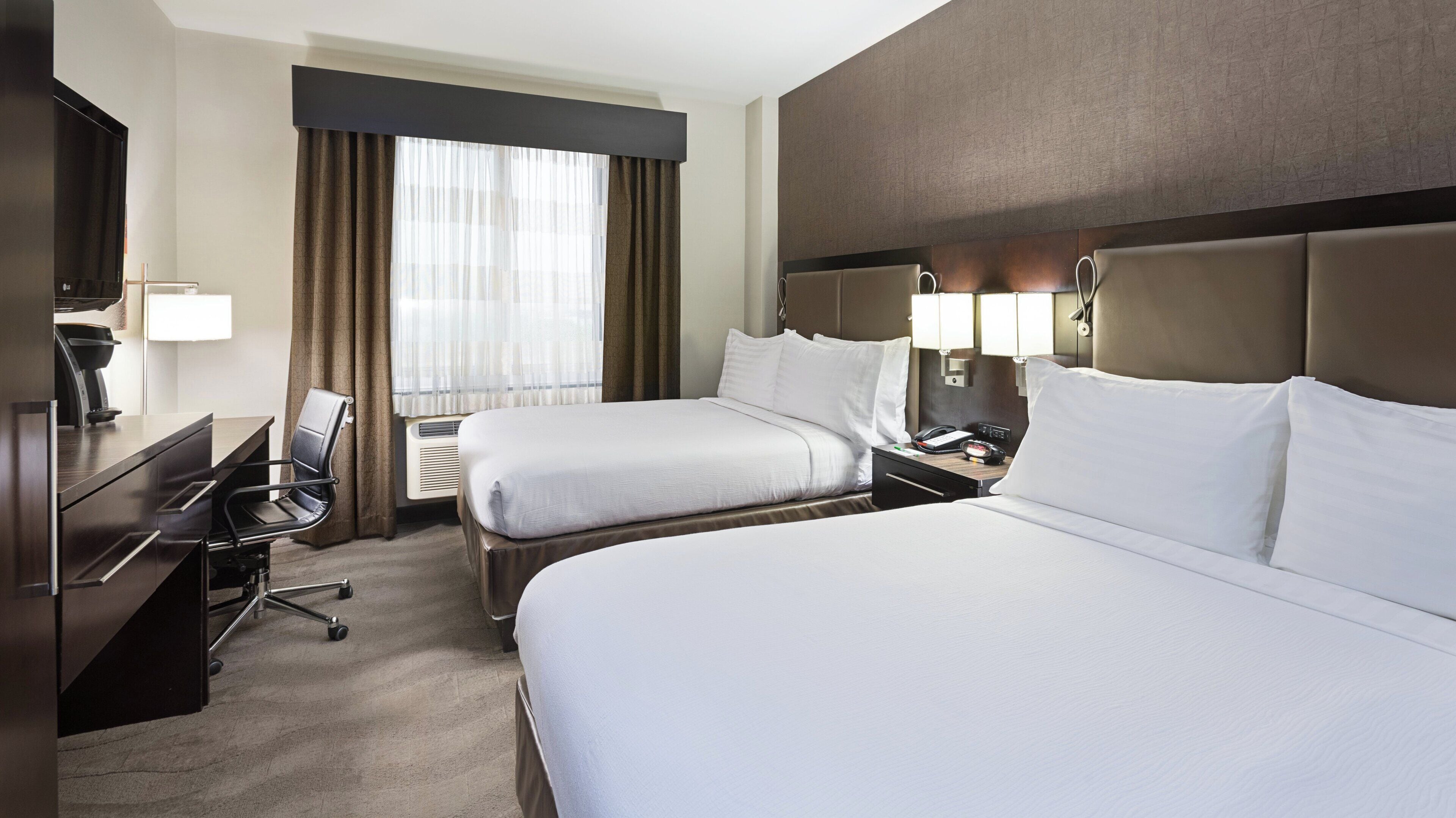 A hotel room with two beds, a desk, a chair, a lamp, and a window with curtains.