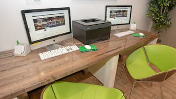 A reception desk with two monitors, a printer, a keyboard, and a mouse. There are two green chairs and a potted plant.