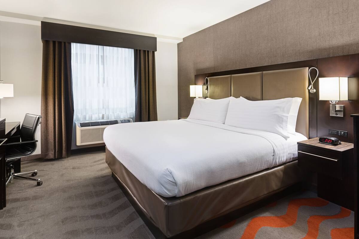 A hotel room with a large bed, a desk with a chair, a TV, and a window with curtains.