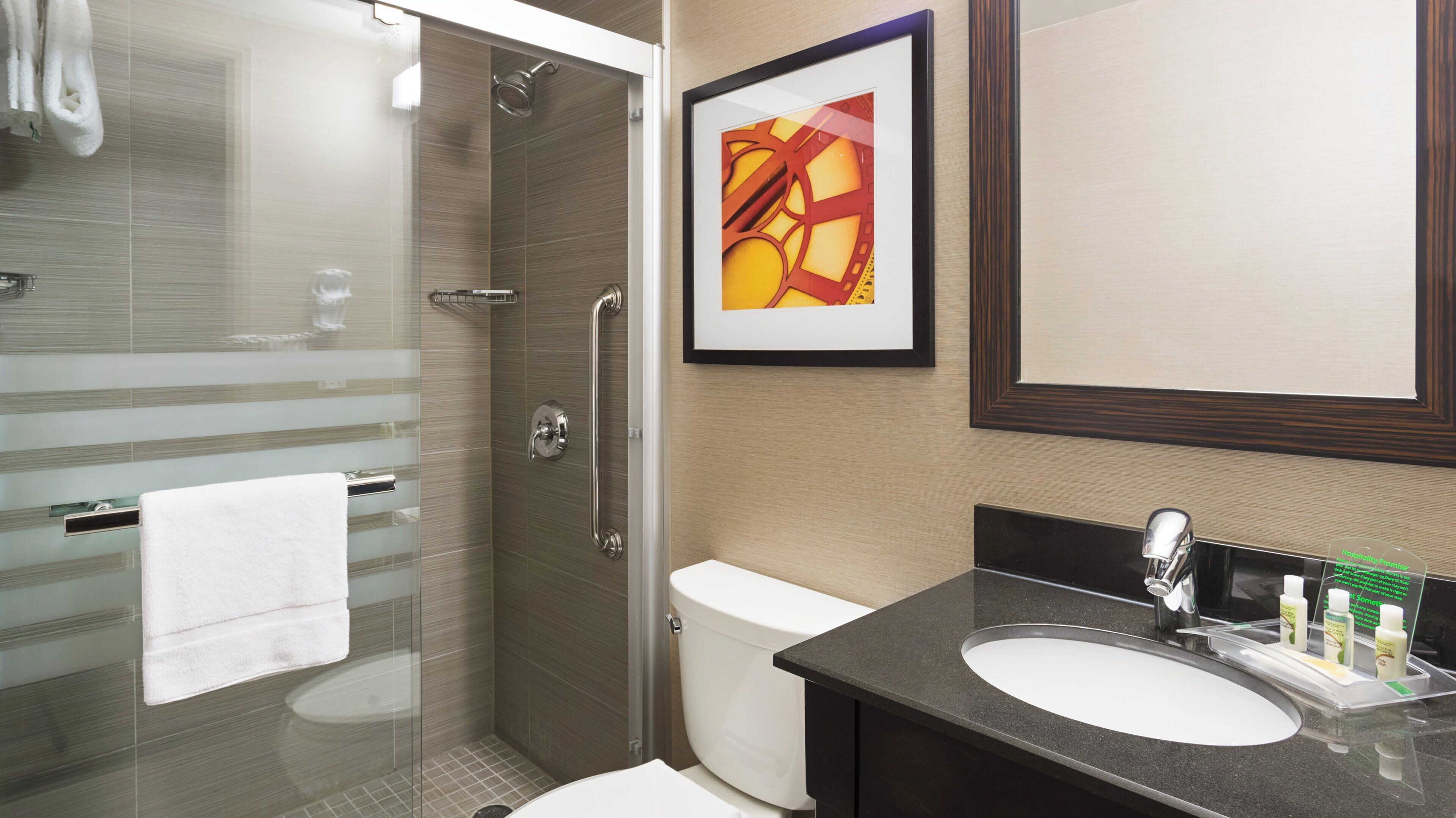 Un bagno moderno con box doccia in vetro, un water bianco, un lavandino con specchio e un quadro astratto appeso alla parete.