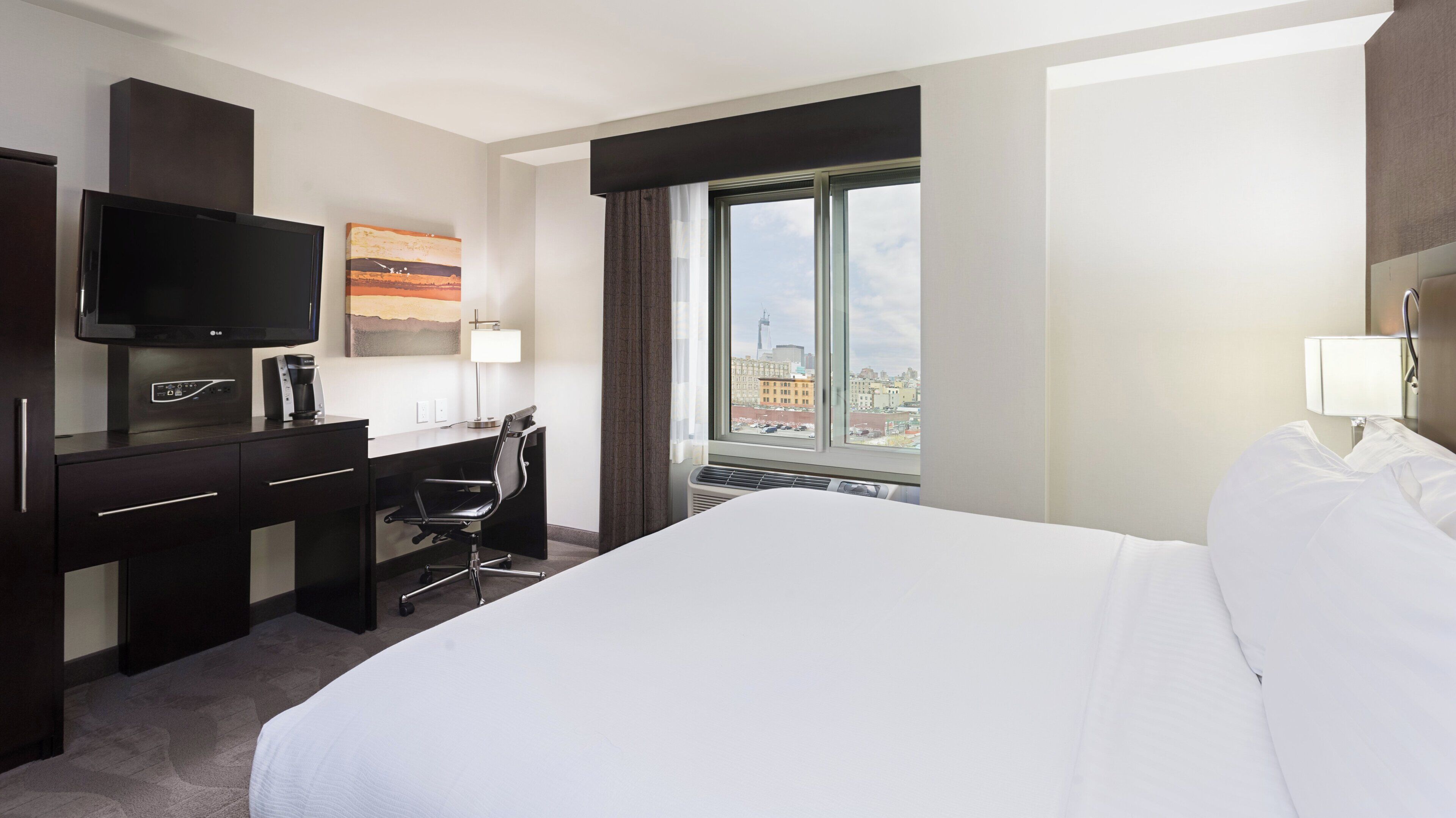 Une chambre d’hôtel équipée d’un lit, d’un bureau, d’une chaise, d’une télévision et d’une fenêtre donnant sur la ville.