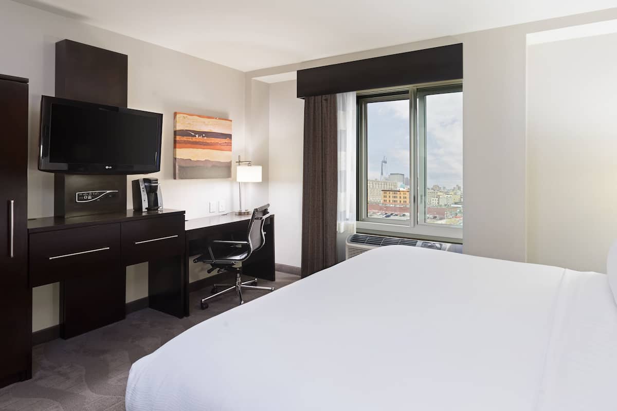 A hotel room with a bed, desk, chair, TV, and a window with a city view.