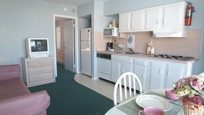A compact kitchen with white cabinets, a microwave, and a stove. There is a dining table with pink cups and a vase of flowers. - The Caprice Motel (Wildwood)