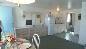 A compact kitchen with white appliances, a dining area with pink chairs and a vase of flowers, and a small TV on the counter. - The Caprice Motel (Wildwood)