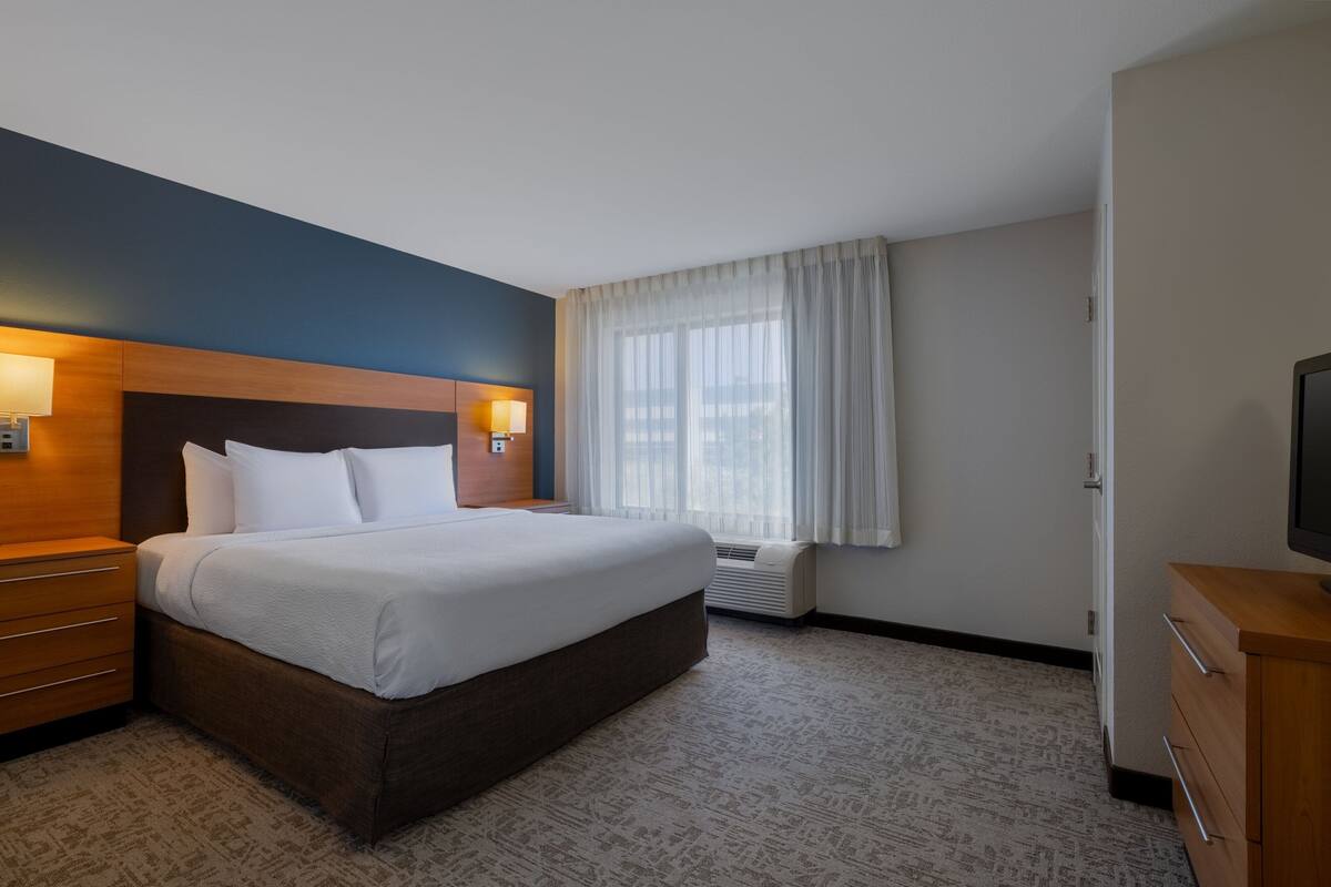 A hotel room with a large bed, a wooden headboard, a nightstand with a lamp, a television, and a window with curtains.
