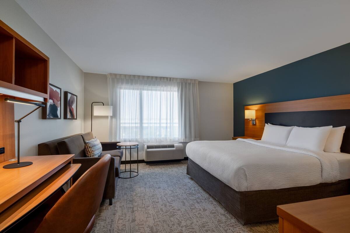 A hotel room with a bed, a desk, a chair, a lamp, and a small table.