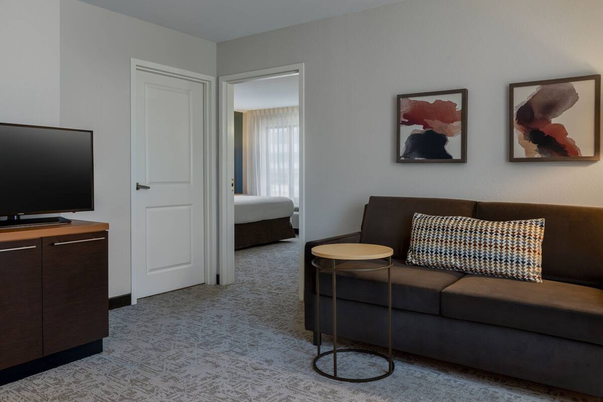 A hotel room with a dark brown sofa, a small round side table, a flat-screen TV on a wooden cabinet, and abstract wall art.