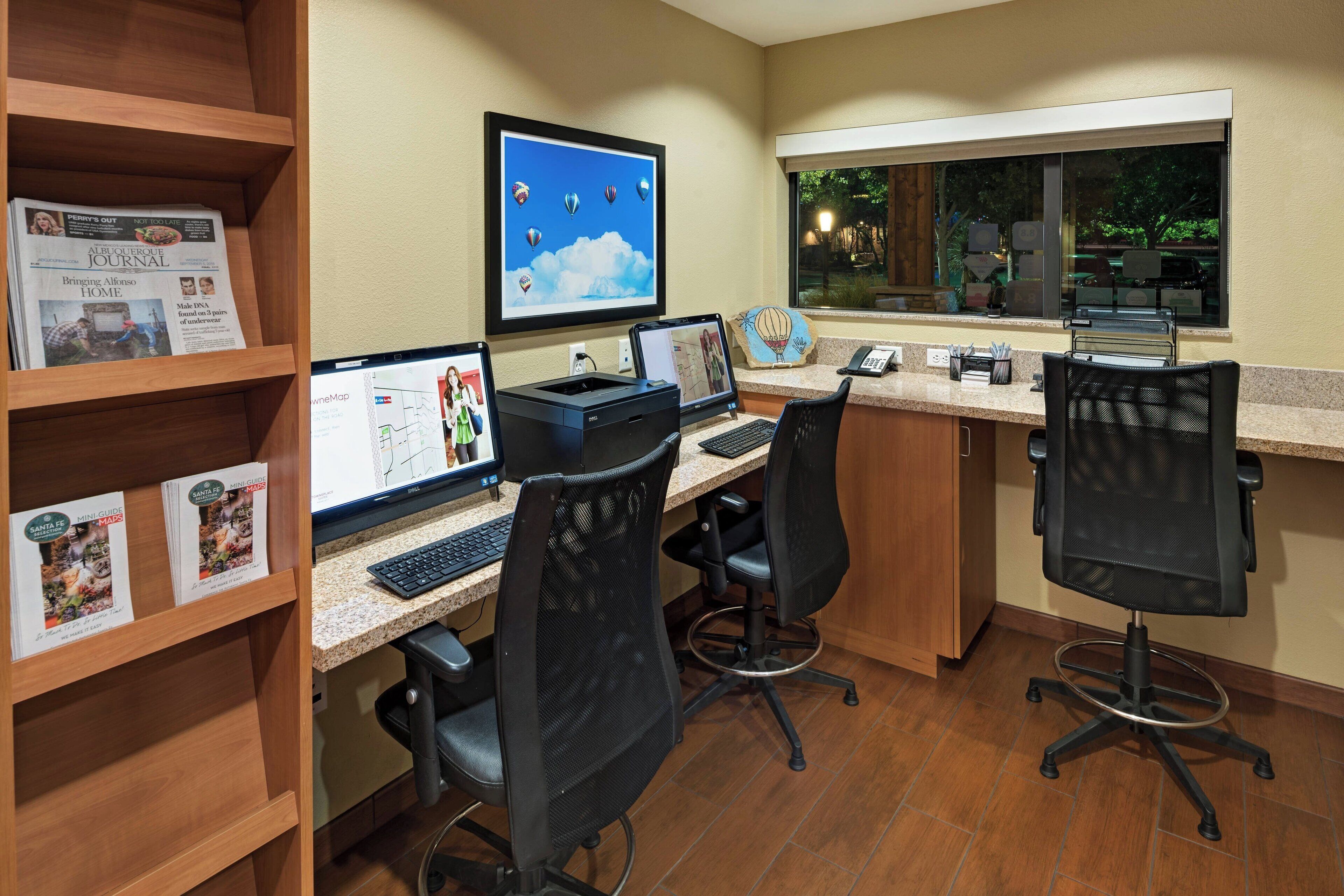 A workspace with two desks, computers, chairs, a window with a view, and a shelf with books.