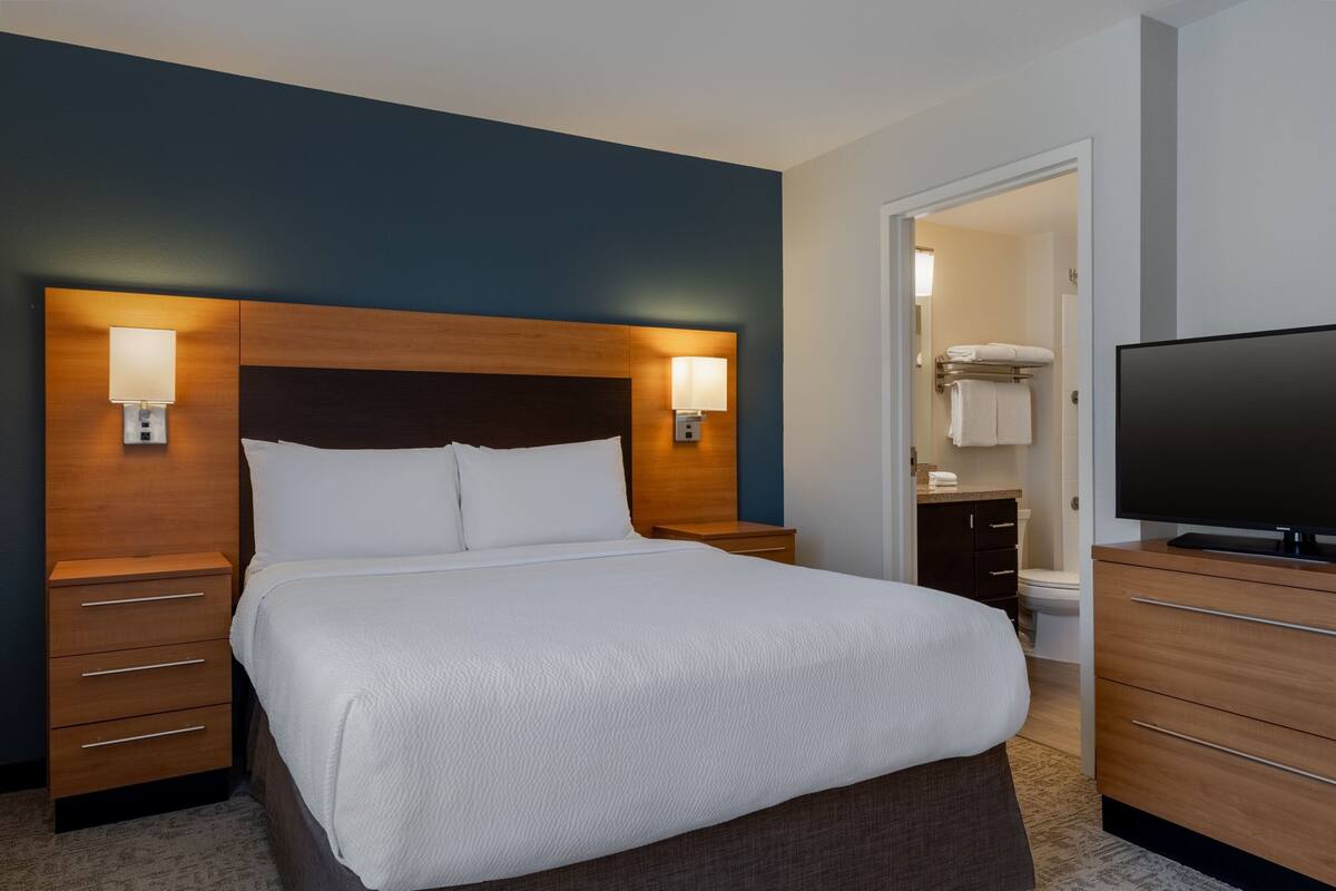 A hotel room with a large bed, bedside lamps, a television, a bathroom with a toilet and sink, and a door leading to another room.